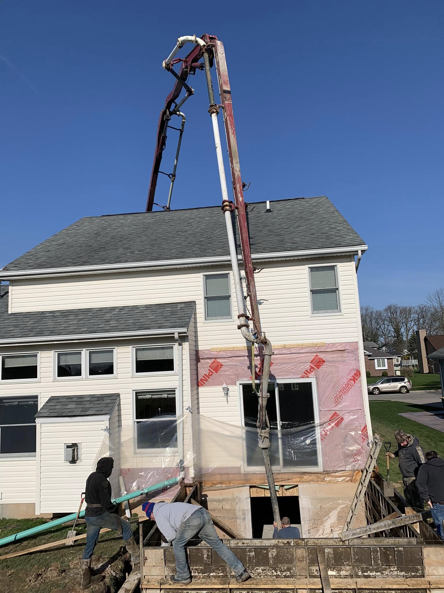 Crew working on foundation expansion for a home addition, with concrete pump hose and excavated area visible.