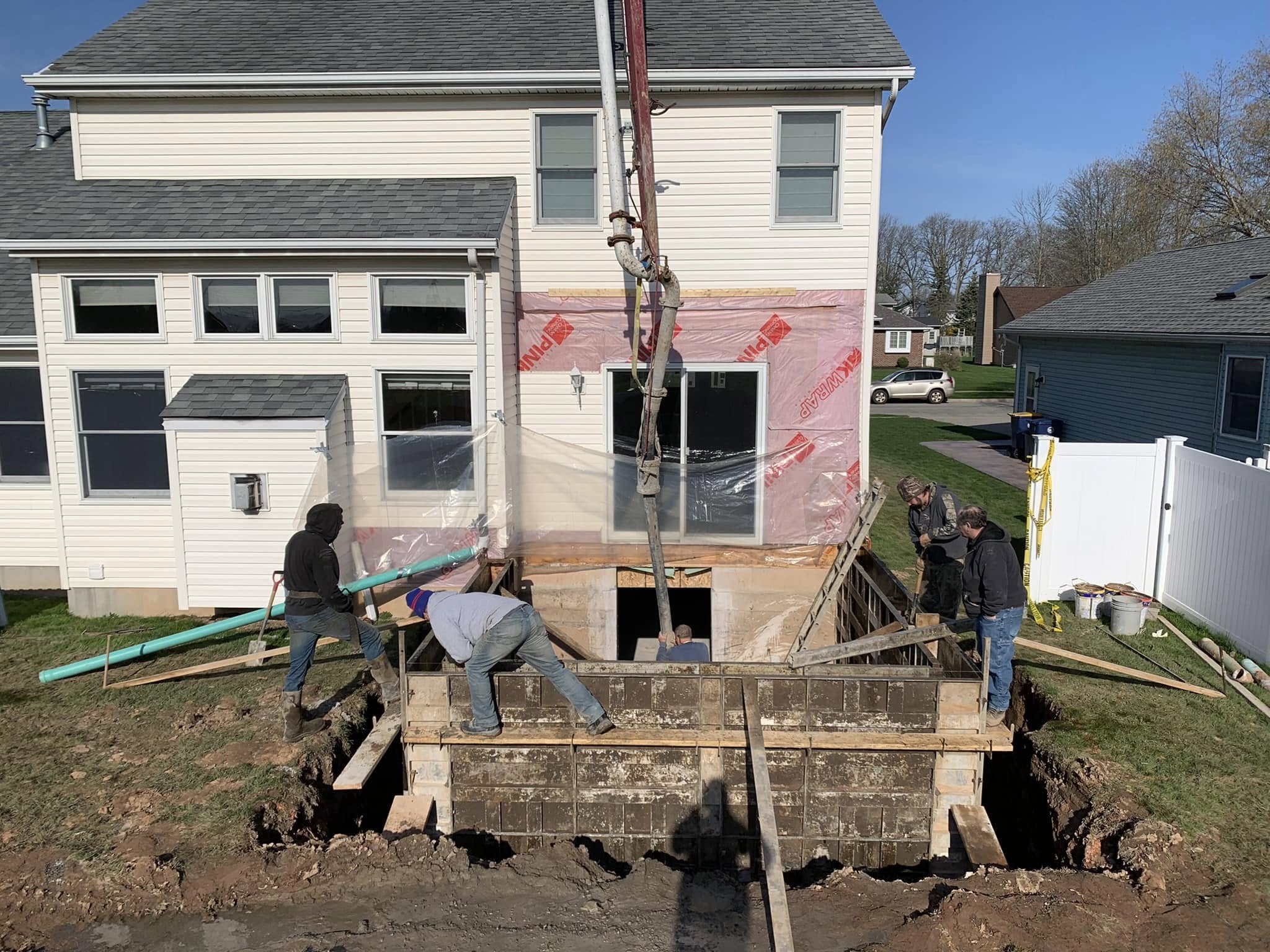 Workers pouring concrete into foundation forms for a home addition, using a pump hose beside an existing house.