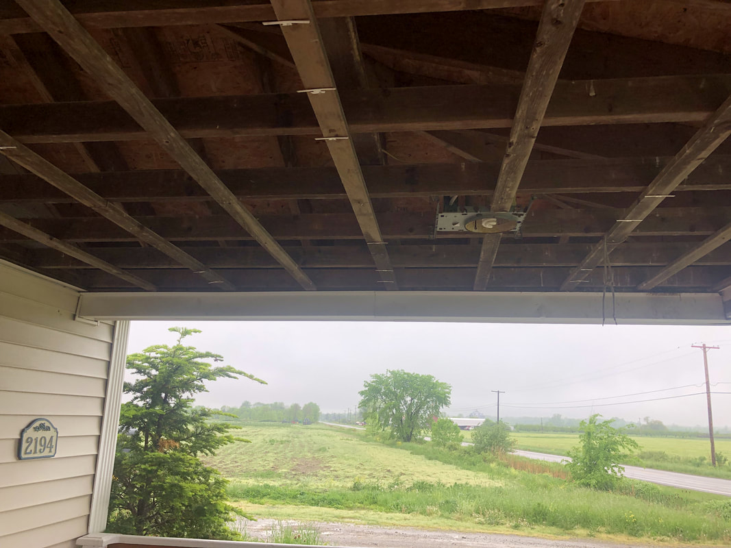 View from beneath partially open porch roof showing exposed beams and rural landscape during ceiling repair.