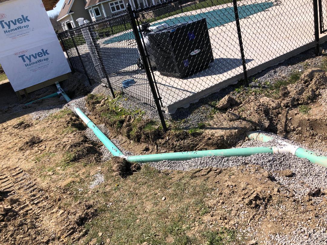 Drainage and plumbing pipes installed in gravel trench beside Tyvek‑wrapped structure near pool equipment.