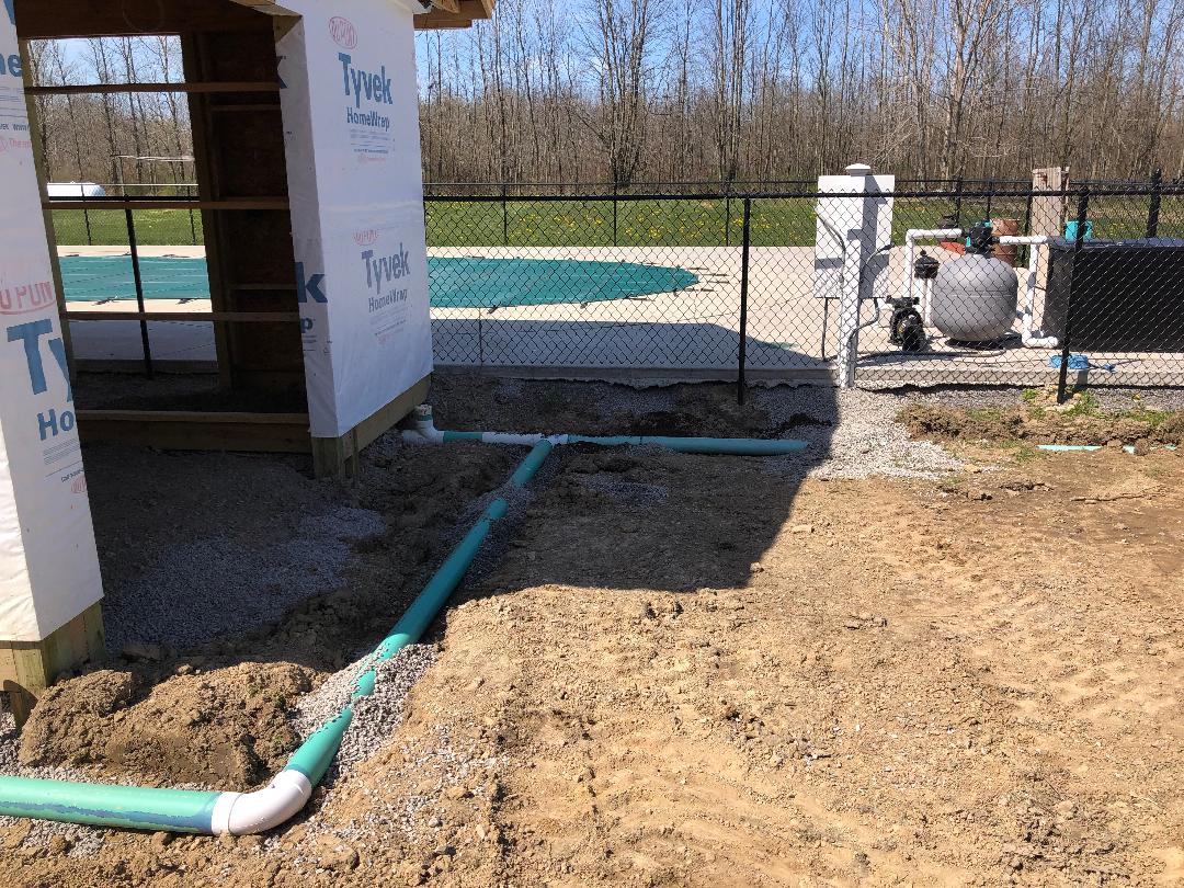 Green PVC drainage pipes forming T‑junction in gravel trench near outdoor utility equipment.